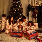 A family of five in matching Small and Big Folk pajamas sits on a white shag rug in a cozy living room, opening Christmas presents in front of a decorated Christmas tree and fireplace mantel with stockings.
