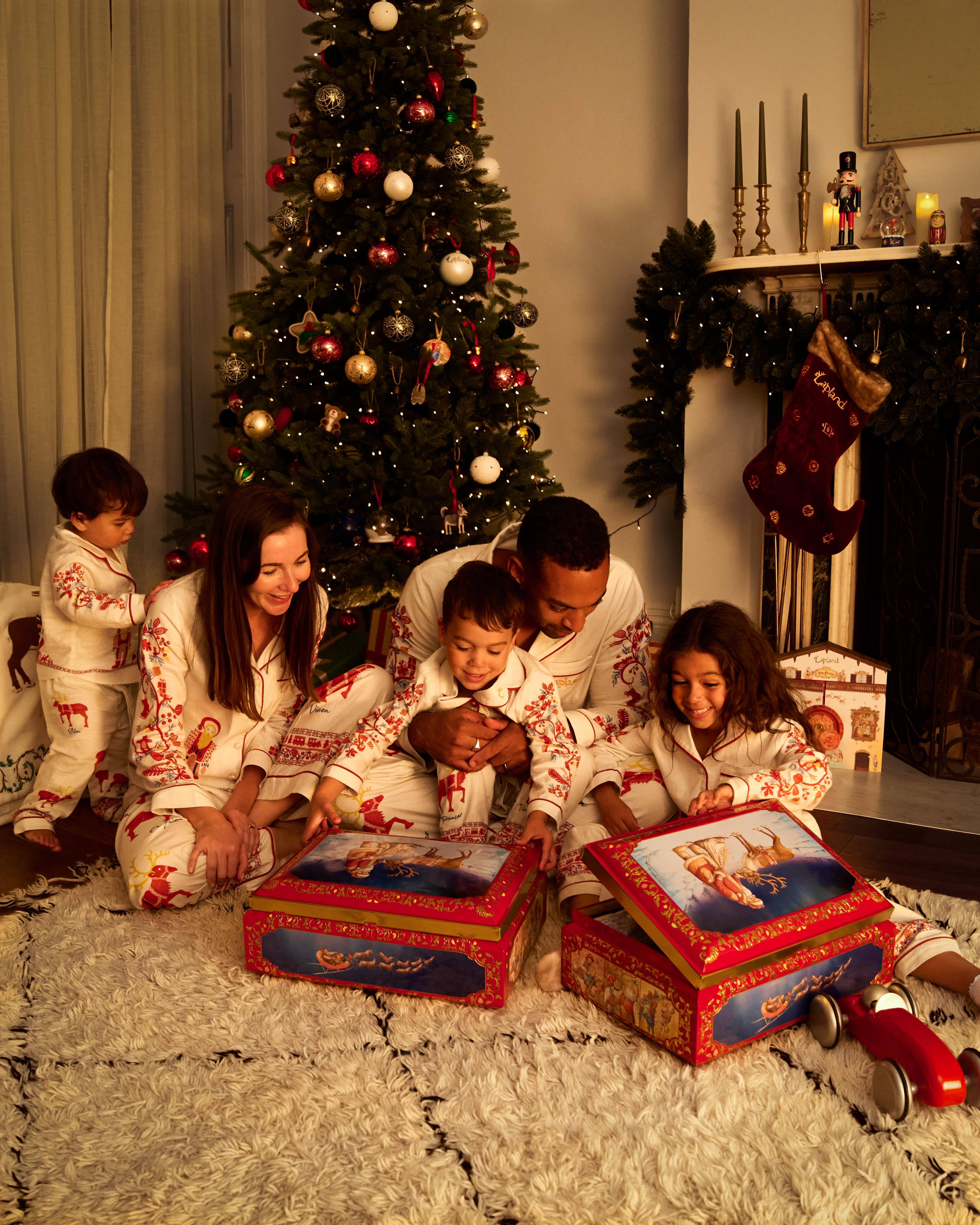 A family of five in matching Small and Big Folk pajamas sits on a white shag rug in a cozy living room, opening Christmas presents in front of a decorated Christmas tree and fireplace mantel with stockings.