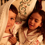 Two children wearing matching holiday-themed Small Folk white pajamas with red trim and floral embroidery, nestled under a hand embroidered cream blanket in a warm, festive indoor setting.