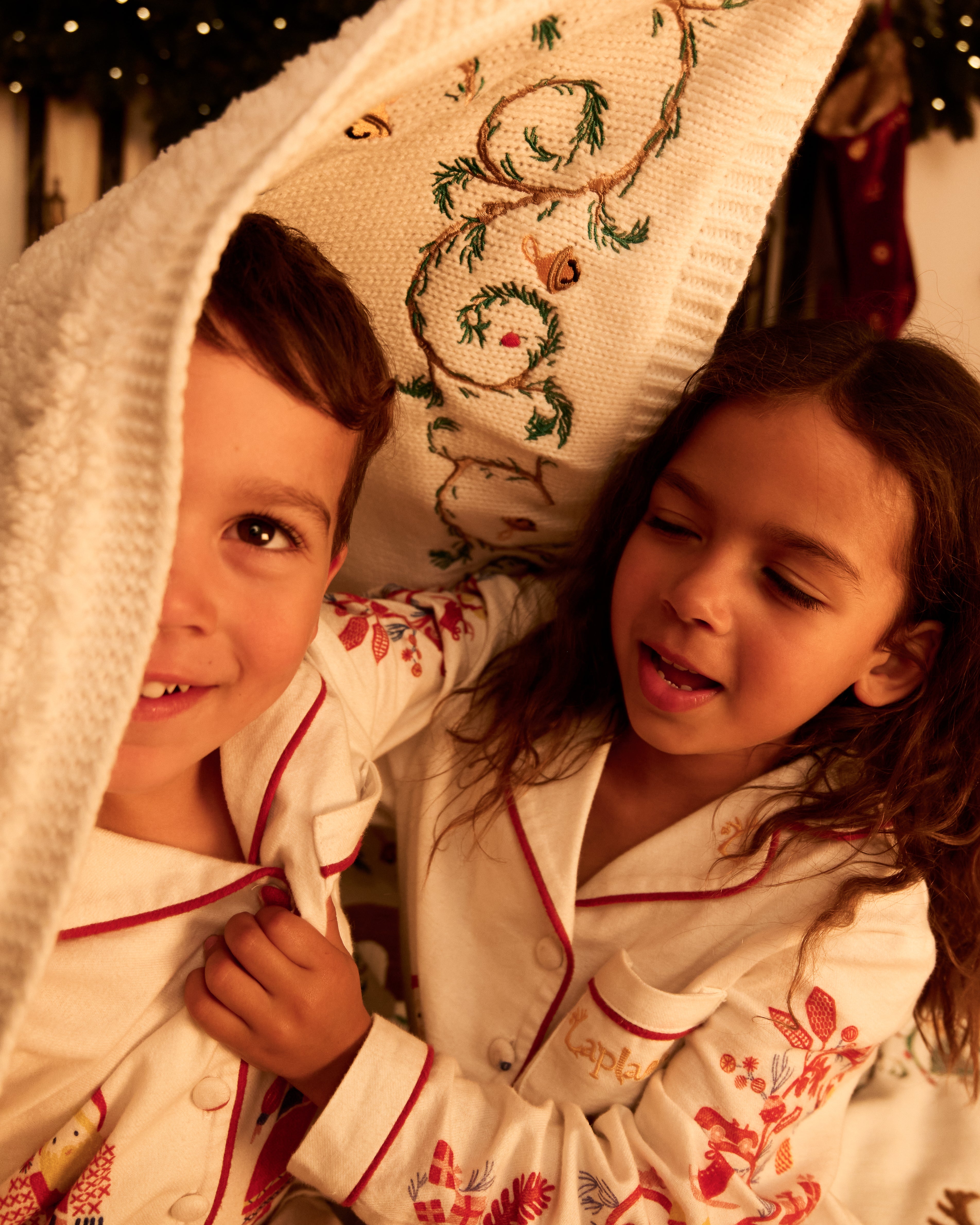 Two children wearing matching holiday-themed Small Folk white pajamas with red trim and floral embroidery, nestled under a hand embroidered cream blanket in a warm, festive indoor setting.