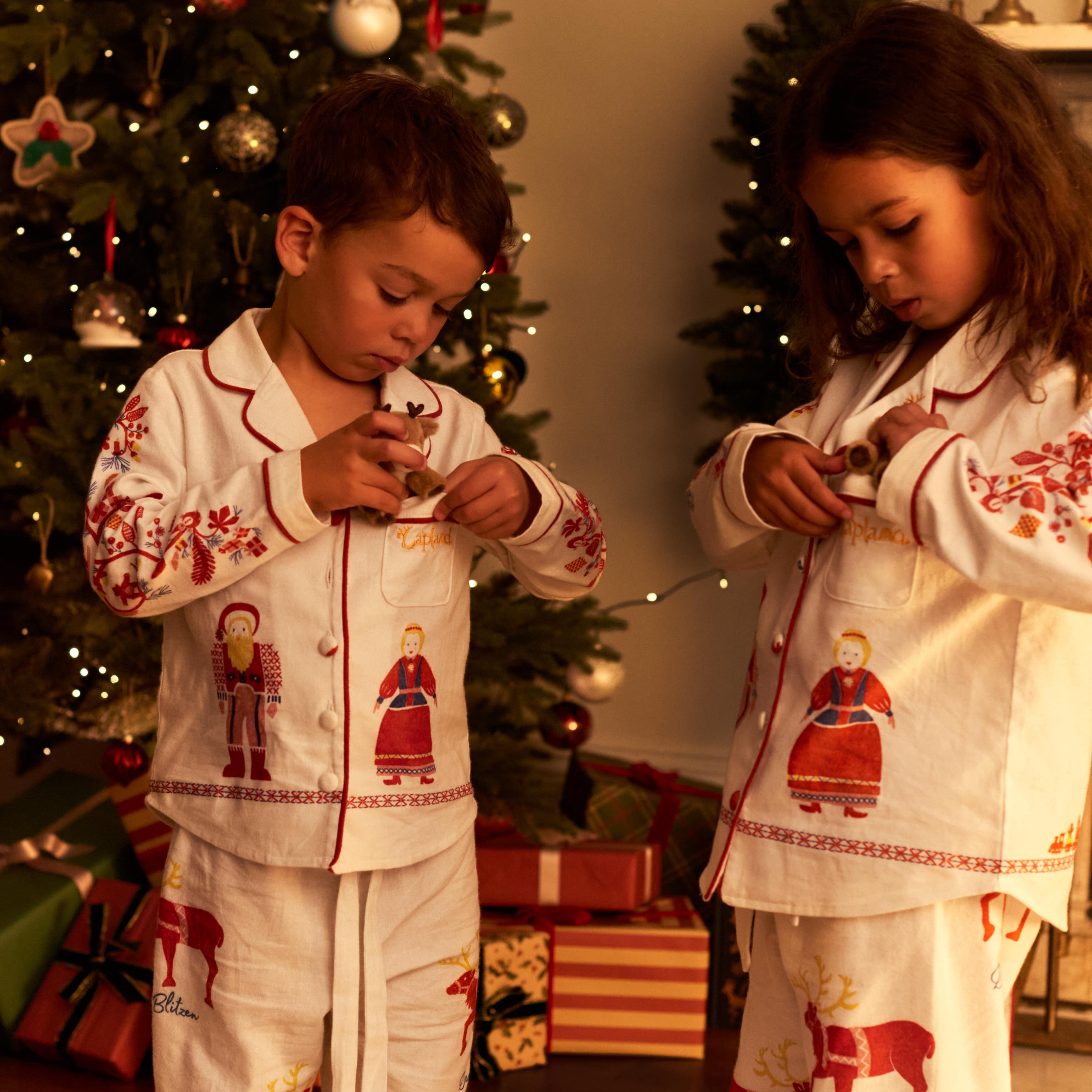 Two children wearing matching Small Folk pajamas standing in front of a decorated Christmas tree in a warmly lit room.