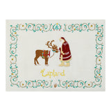 A square white hand embroidered blanket featuring a Christmas scene with Father Christmas and a brown reindeer, surrounded by a decorative teal and gold border. The word 'Lapland' is embroidered in gold near the bottom center.