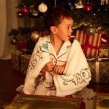 A young boy sits on the floor in front of a decorated Christmas tree, wrapped in the Lapland blanket with red trim and green embroidered holiday motifs. Gift boxes are wrapped around the tree, and a gold Lapland Christmas Eve Chest is in the foreground.