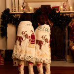 Two children standing with their backs to the camera, wrapped in matching Lapland Christmas blankets and wearing matching Small Folk pajamas with Father Christmas and reindeer designs. The scene is set in a cozy, decorated holiday living room in front of a fireplace.
