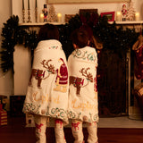 Two children standing with their backs to the camera, wrapped in matching Lapland Christmas blankets and wearing matching Small Folk pajamas with Father Christmas and reindeer designs. The scene is set in a cozy, decorated holiday living room in front of a fireplace.