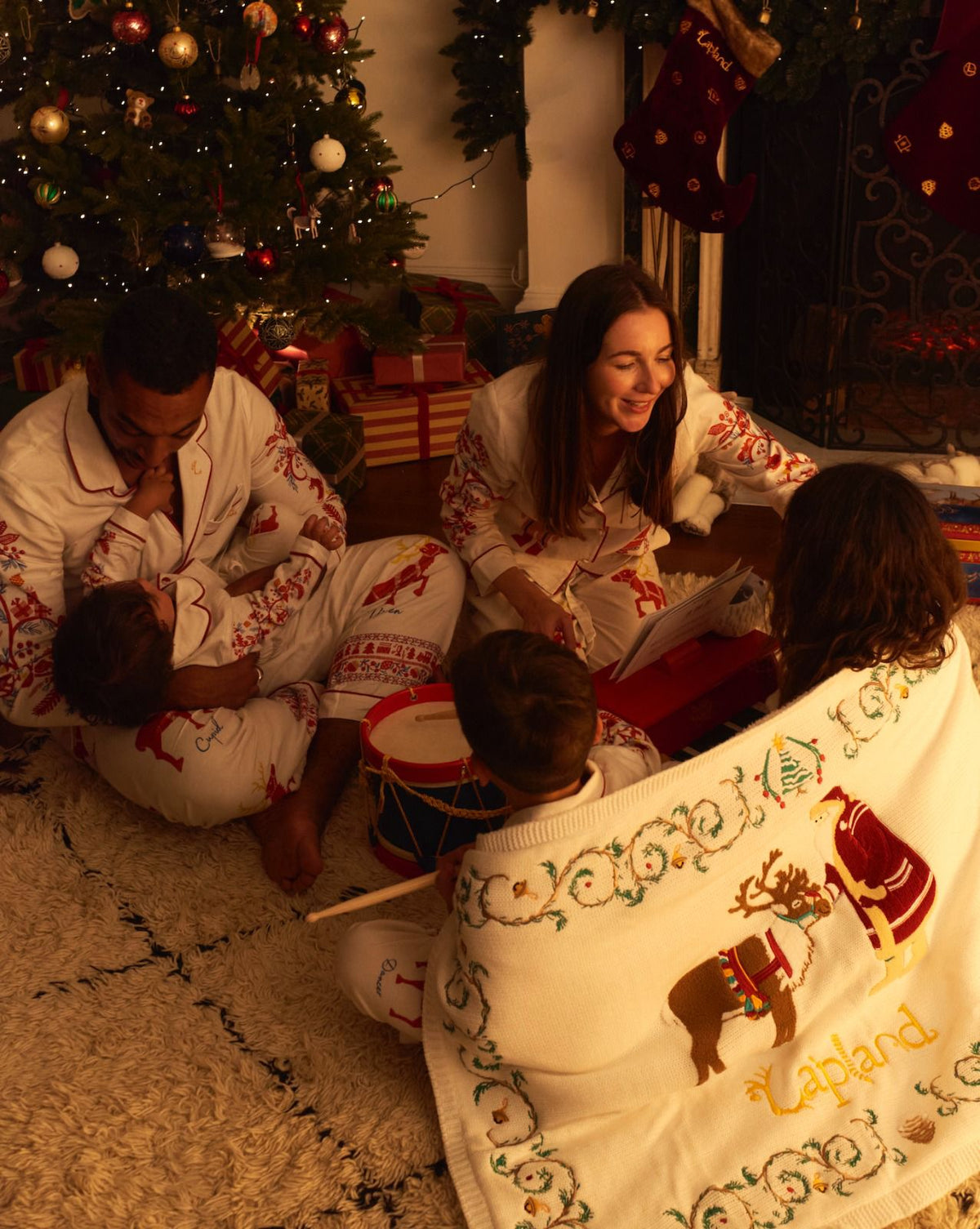 A family of four in matching Small & Big Folk Christmas pajamas gathered on a rug in front of a decorated Christmas tree, with a white embroidered Lapland blanket featuring Father Christmas and a reindeer in the foreground.